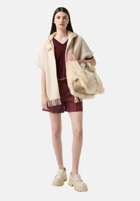 Béžový pončo s třásněmi, vínový top, burgundské kraťasy a krémové platformové boty. Drží tašku s umělou kožešinou. Hladké textury.