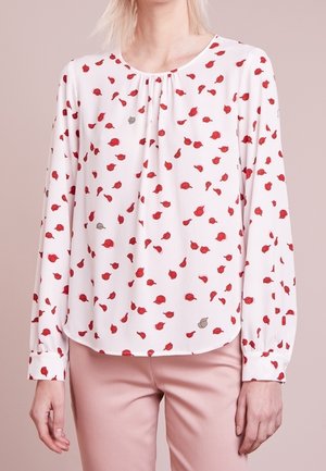 Blouse blanche à manches longues avec petit motif floral rouge et gris portée avec un pantalon rose clair sur une personne devant un fond neutre.