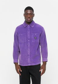 Purple cotton shirt with long sleeves, button-up front, two chest pockets, and a small logo patch. Soft texture and relaxed fit.