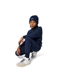 Fato de treino azul-marinho com jaqueta de fecho éclair e punhos elásticos, combinado com um gorro de malha a condizer. Ténis altos brancos e cinzentos com atacadores pretos.