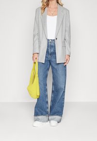 Kvinna som bär en grå blazer, vit topp, vida blå jeans med uppvikta ben, vita sneakers och håller en ljusgul handväska vid sin sida.