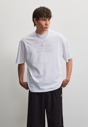 Jeune homme aux cheveux courts et foncés portant un T-shirt blanc avec l’inscription « Ma copine est plus sexy que la tienne » et un pantalon noir, les mains dans les poches.