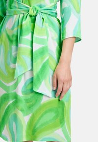 Grün-weißes, abstrakt gemustertes Kleid mit einer gebundenen Taillenschleife, drei Viertelärmeln und einer sichtbaren Hand neben dem Kleid.