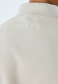 Polo shirt beige chiaro con tessuto testurizzato, dotato di colletto corto e logo "PXP" traslucido sul retro.