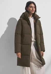 Manteau rembourré vert olive avec capuche, fermeture éclair à l'avant et sections matelassées. Porté sur un sweat-shirt léger et un pantalon ample, avec une texture lisse.