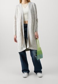 Hellgraue, gestrickte Strickjacke mit offenem Vorderteil, Kabelmuster und langen Ärmeln; kombiniert mit hochtaillierten blauen Jeans und einer grünen Farbverlauf-Tasche.