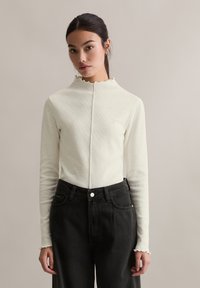 Kremowy prążkowany top z długim rękawem i wysokim, karbowanym dekoltem. W połączeniu z czarnymi jeansami z wysokim stanem i zapięciem na guziki z przodu.