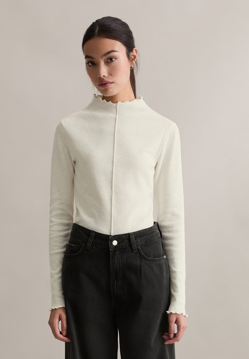 Kremowy prążkowany top z długim rękawem i wysokim, karbowanym dekoltem. W połączeniu z czarnymi jeansami z wysokim stanem i zapięciem na guziki z przodu.