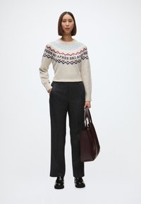 Kootud sviiter kreemja alusega, värviline geomeetriline muster sinise, punase ja mustaga, kombineeritud hallide täpsetega pükstega ning burgundi värvi kätetaskuga.