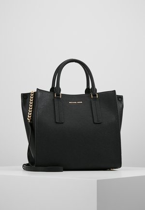 Schwarze, genarbte Lederhandtasche von Michael Kors mit doppelten Griffen, goldener Kettenriemen und minimalistischem goldenen Logo auf der Vorderseite, auf einer weißen Oberfläche platziert.
