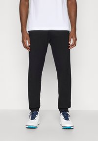 Svarta sweatpants med avsmalnande ben, elastiska muddar och en mjuk yta. Bärs med vita sportskor som har blå accenter.