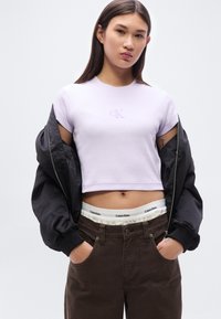 Μωβ κοντοκομμένο μπλουζάκι με κεντημένο λογότυπο, συνδυασμένο με σκούρο καφέ παντελόνι που έχει εμφανή μέση. Μαύρο μπουφάν bomber ριγμένο στους ώμους.