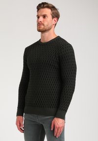 Suéter de punto verde oscuro con un patrón texturizado, cuello redondo y dobladillos y puños acanalados. Combinado con jeans grises.