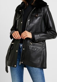 Chaqueta de cuero negra con cuello de piel, cierre de cremallera y botones a presión, dos bolsillos en el pecho y un bolsillo lateral con cremallera, textura suave y ajuste entallado.