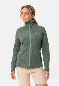 Giacca in pile verde scuro con una trama a motivi; presenta un colletto alto e una cerniera a contrasto. Indossata con pantaloni corti di colore chiaro.