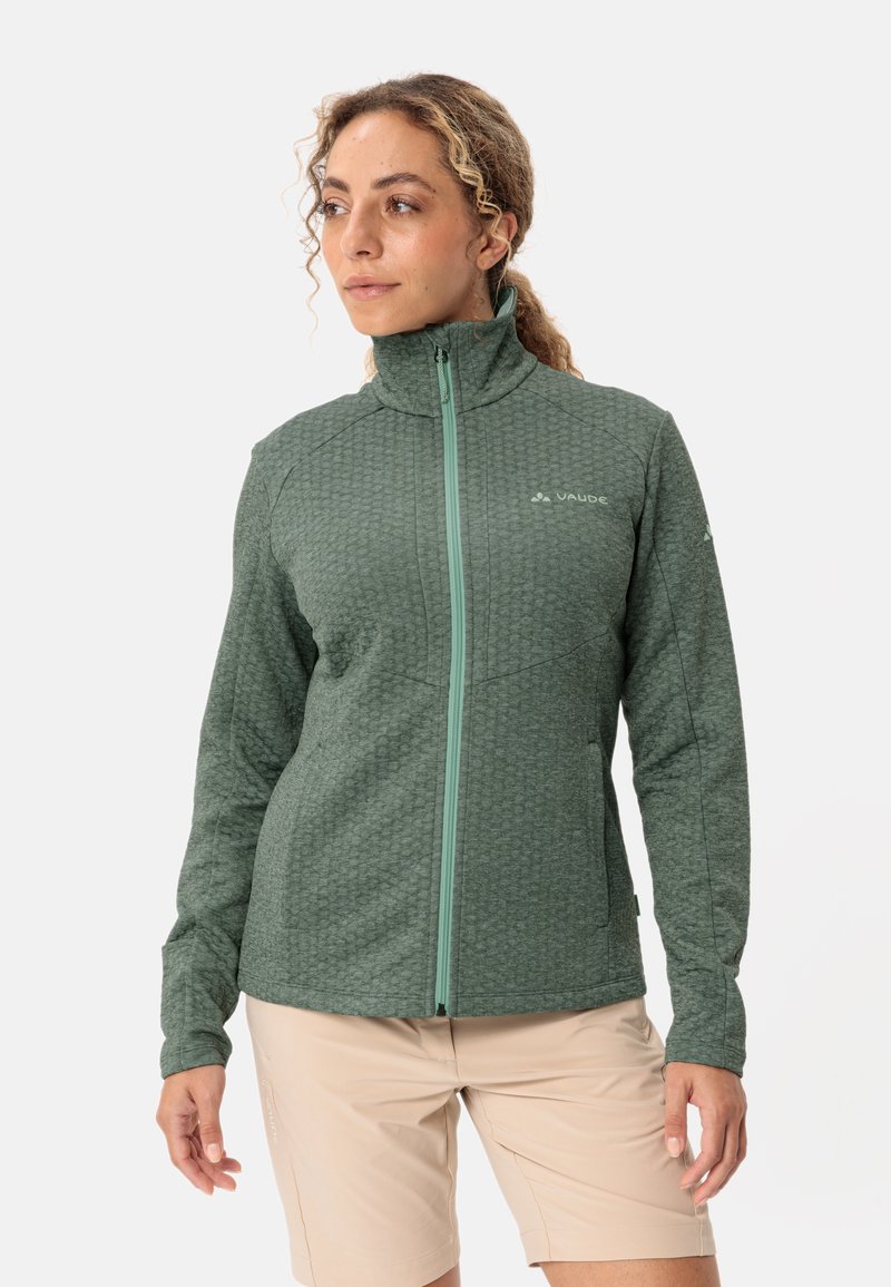 Giacca in pile verde scuro con una trama a motivi; presenta un colletto alto e una cerniera a contrasto. Indossata con pantaloni corti di colore chiaro.