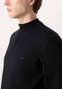 Pulover negru, tricotat, cu fermoar la jumătate și guler înalt. Prezintă un logo subtil "BOSS" pe piept. Textură moale, design care se mulează pe corp.