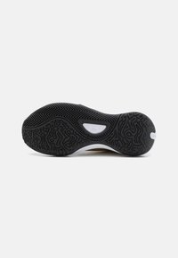 Semelle d'une chaussure de sport noire avec une bande de roulement texturée, un motif en chevrons et une insertion de semelle intermédiaire blanche visible au niveau de la voûte plantaire.