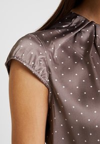 Bruine satijnen blouse met witte stippen, ronde kraag en korte pofmouwen. Gladde textuur met geplooide stofdetails op de schouders.