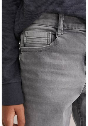 Jean en denim gris avec une texture lisse, comportant un design de poche arrière et des détails à la ceinture. Porté avec un haut à manches longues côtelé de couleur sombre.