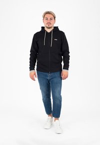 Schwarze Zip-Up-Hoodie mit beigen Kordeln, mit einem Logo auf der Brust. Getragen mit blauen Jeans und weißen Sneakers. Einfarbig, lässiger Schnitt.