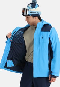Junger Mann in blauer Winterjacke und gemustertem Beanie, der die Innenmerkmale und Etiketten der Jacke zeigt, mit Skibrille auf dem Beanie.