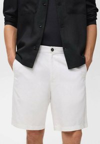 Pantalones cortos de algodón blancos con un corte recto, que presentan un cierre de botones en la parte frontal y bolsillos laterales, combinados con una camisa negra.