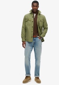 Zielona kurtka na brązowym swetrze, jasnoniebieskie dżinsy z wyblakłymi miejscami i beżowe zamszowe buty. Zegarek widoczny na nadgarstku.