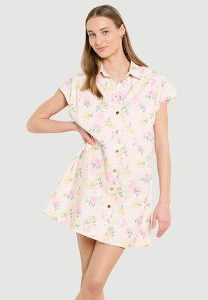 Junge Frau steht mit einer Hand auf der Hüfte und trägt ein kurzärmliges, vorne mit Knöpfen versehenes Blumenkleid in Pastellrosa, Gelb und Grün auf cremefarbenem Hintergrund.