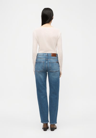 Γυναίκα με μακριά σκούρα μαλλιά, που φοράει μακρυμάνικη μπεζ μπλούζα, τζιν ψηλόμεσο σε μπλε απόχρωση και μαύρα παπούτσια με τακούνι, στέκεται γυρισμένη με την πλάτη.