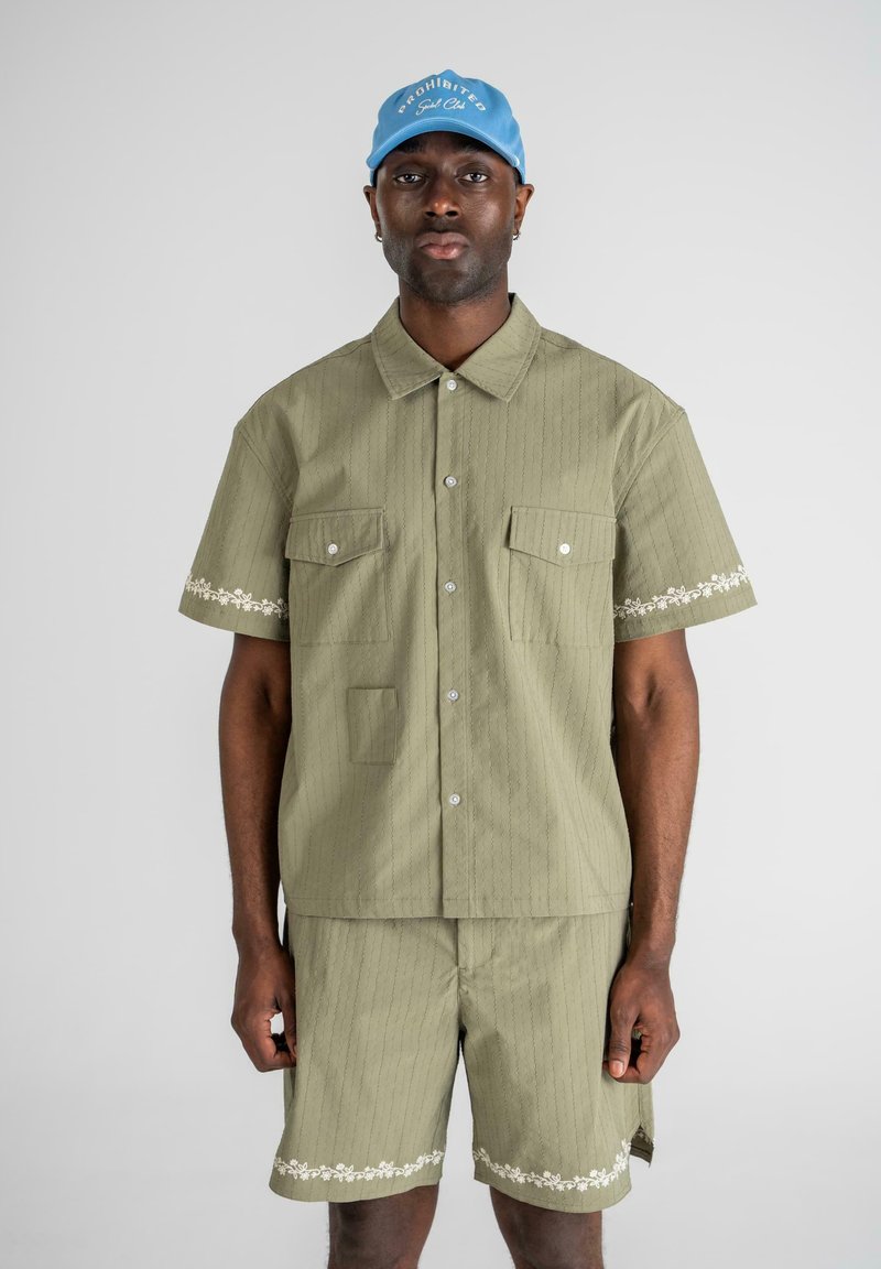 Oliven grønn kortermet skjorte og matchende shorts, med vertikale striper og blomsterdetaljer på mansjettene og kantene. Blå caps på hodet.