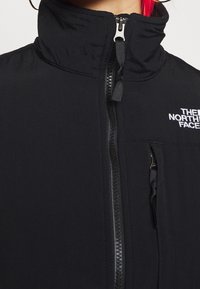 Svart zip-up jacka med hög krage, som har en framsida med dragkedja, en broderad logotyp och en slät, matt textur.