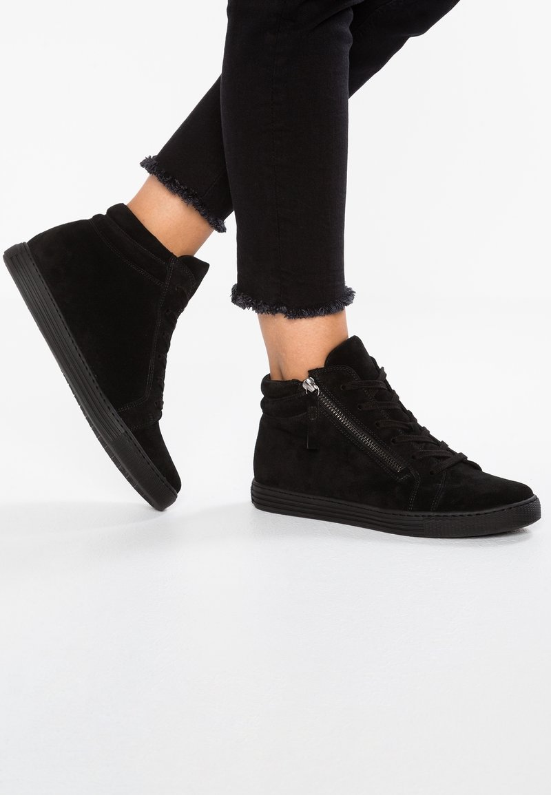 Pieds portant des baskets montantes en daim noir avec des fermetures éclair latérales et des lacets à l'avant, associés à un jean skinny noir à ourlet effiloché.