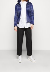 Blå puffjakke med vandret syninger, lynlås og hætte; parret med en hvid skjorte med knapper og løse sorte bukser. Hvide sneakers.