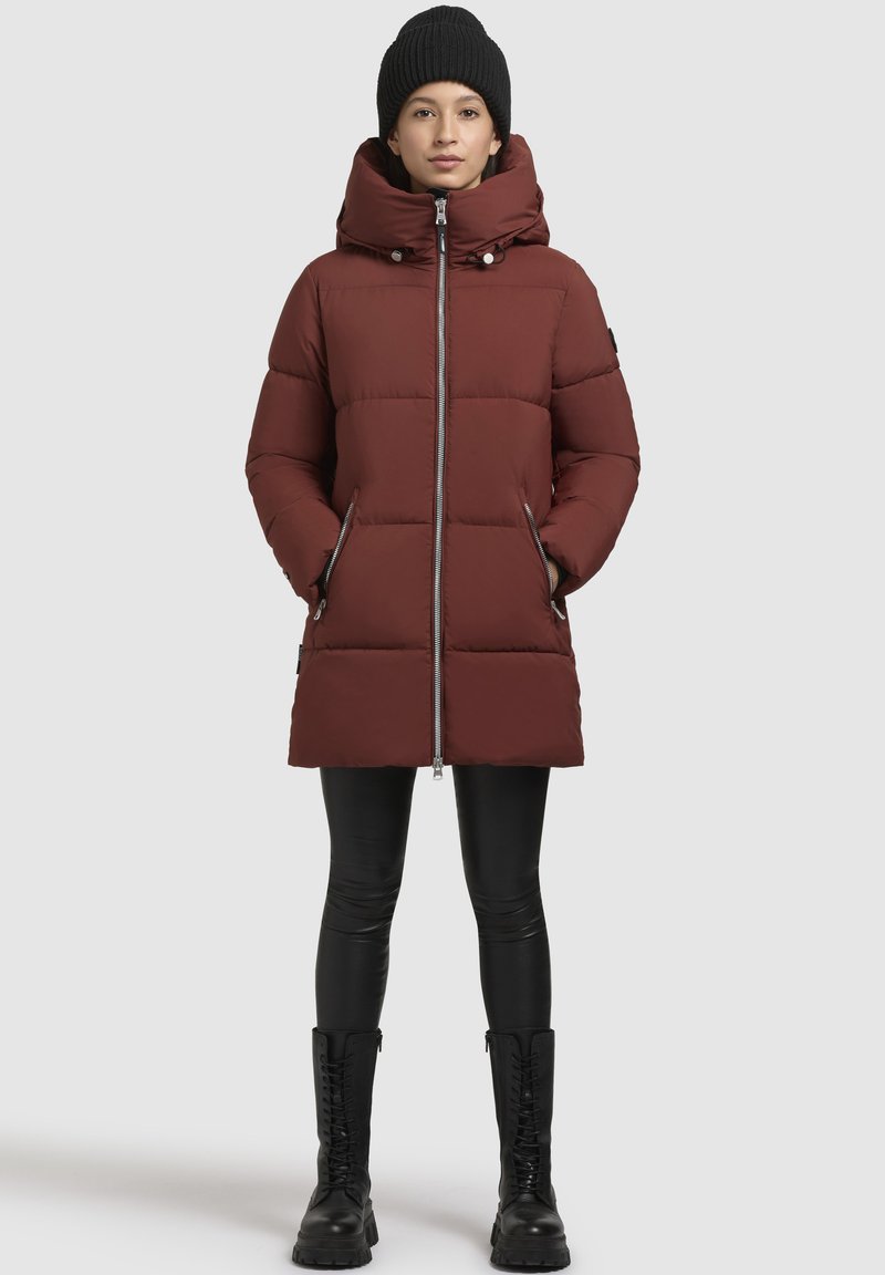 Bordeaux pufferjakke med høj krave, lynlås lukning og to sidelommer; båret med sorte leggings og knæhøje snørebåndsstøvler.