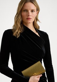 Černé sametové šaty s obálkovým designem, dlouhými rukávy. Zlatá kožená kabelka s embosovaným logem, držena v pravé ruce. Hladká textura, elegantní vzhled.