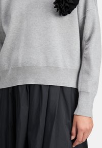 Hellegrauer, gestrickter Pullover mit geripptem Saum und Bündchen, versehen mit einem schwarzen Stoffblumenakzent auf der Vorderseite, kombiniert mit einem schwarzen Rock.