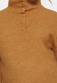 Gros plan sur un pull en tricot couleur rouille avec col montant et cinq boutons recouverts de tissu sur le devant.