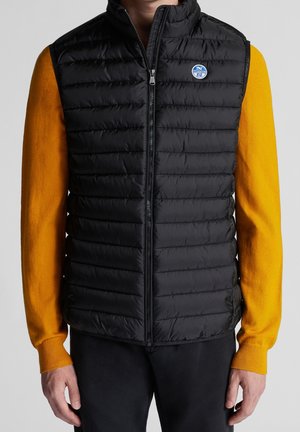 Homme portant un gilet matelassé noir à fermeture éclair devant, par-dessus un pull orange vif à manches longues, debout devant un fond gris uni.