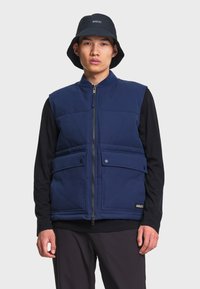 Homme portant un gilet matelassé bleu avec poches avant, une chemise noire à manches longues, un pantalon noir et un chapeau bob noir avec le logo Aigle.