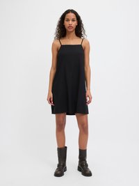 Vestido preto sem mangas com decote quadrado, apresentando um corte solto que termina acima dos joelhos, combinado com botas castanhas até o tornozelo.