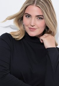 Frau mit blonden Haaren, die silberne Creolen und ein schwarzes Langarmoberteil trägt, sanft lächelnd mit der Hand nah am Gesicht vor einem weißen Hintergrund.