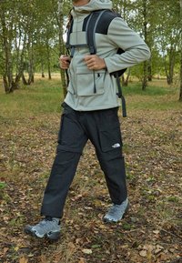 Hellgraue Fleecejacke mit Taschen, schwarze Cargo-Hosen und graue Wanderschuhe, getragen in einem bewaldeten Gebiet mit fallenden Blättern.