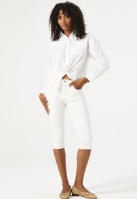 Chemise blanche nouée à la taille, associée à un pantalon blanc cropped. Les chaussures sont en matériau tissé de couleur beige. Le mannequin se tient avec une main sur la hanche.