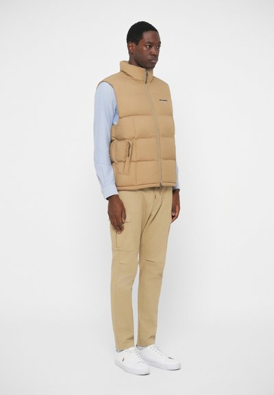Gilet imbottito beige con colletto alto, cerniera frontale e tasche laterali, indossato sopra una camicia azzurro chiaro e abbinato a pantaloni beige e scarpe bianche.