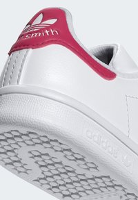 Baskets blanches avec un dessus en cuir texturé, accents roses au niveau du talon et semelle en caoutchouc avec un motif de semelle et une marque.