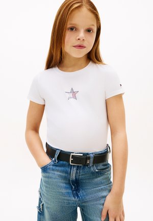 Mladé dievča s dlhými červenými vlasmi, ktoré má na sebe biele tričko s hviezdovým dizajnom a modré džínsy s čiernym opaskom, stojí s jednou rukou v vrecku.