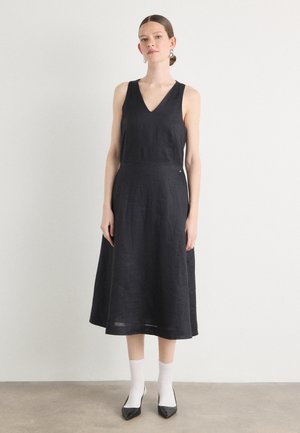Robe en lin noire avec un décolleté en V, sans manches, jupe évasée et ourlet au genou. Le modèle porte des chaussettes blanches et des chaussures noires à bouts pointus.