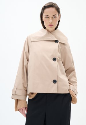 Beige cropped jack met een brede kraag en zwarte knopen, met een gladde textuur. Het model draagt een gebreide trui onder de jas.