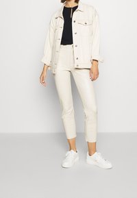 Krémová denimová bunda, černý top, béžové slim fit kalhoty a bílé tenisky. Bunda má knoflíkové zapínání a kapsy, s ohrnutými rukávy.